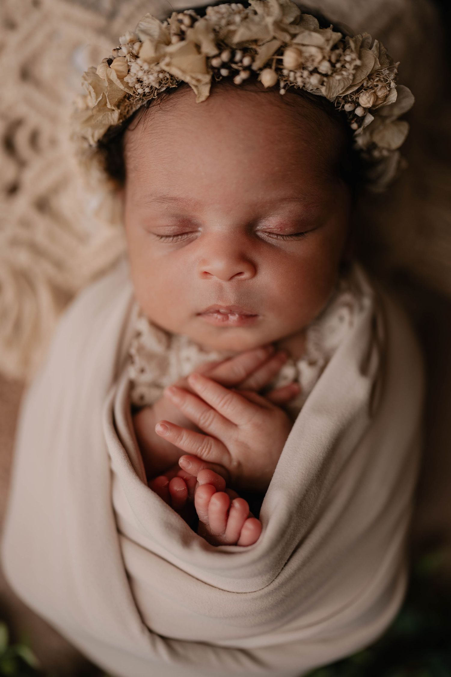 A sleeping newborn baby wearing a delicate floral crown and wrapped in a soft beige swaddle.