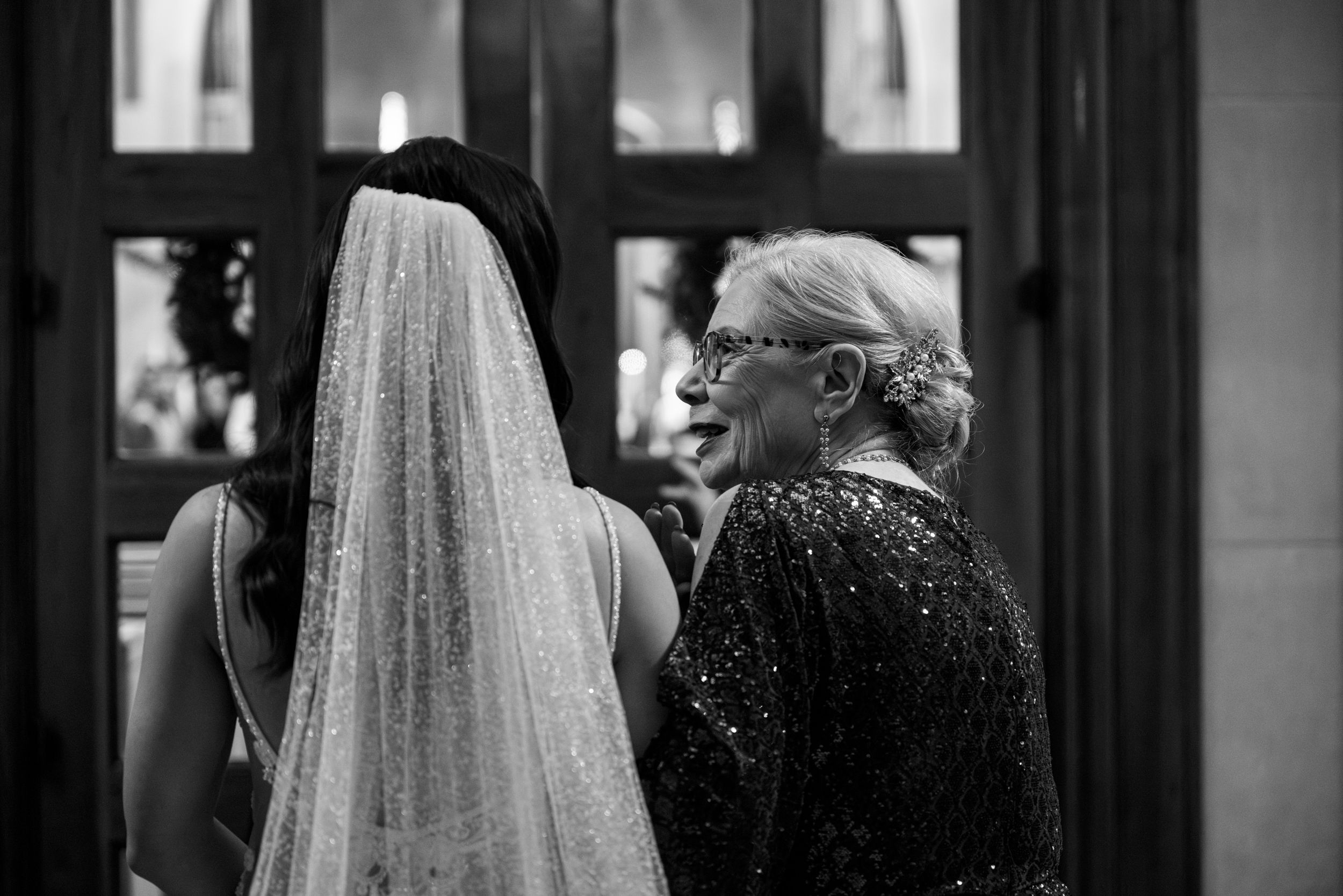 A tender black and white moment between a bride in a veil and woman in sparkly formal wear before a wedding ceremony.