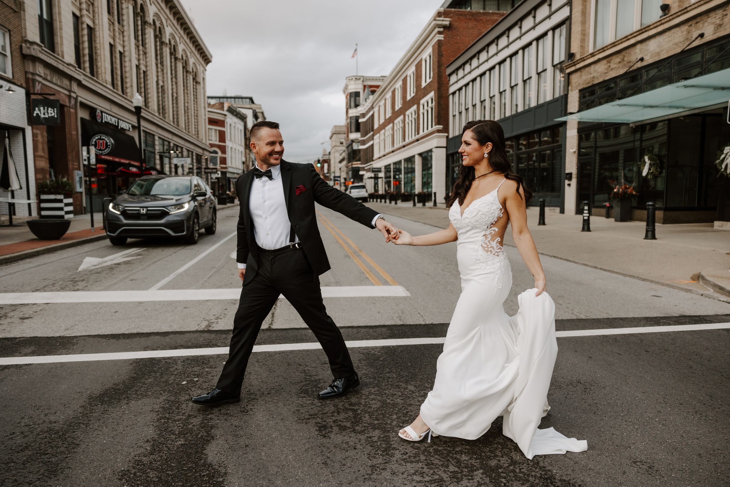 A couple in wedding attire playfully poses in the middle of a downtown street crossing.