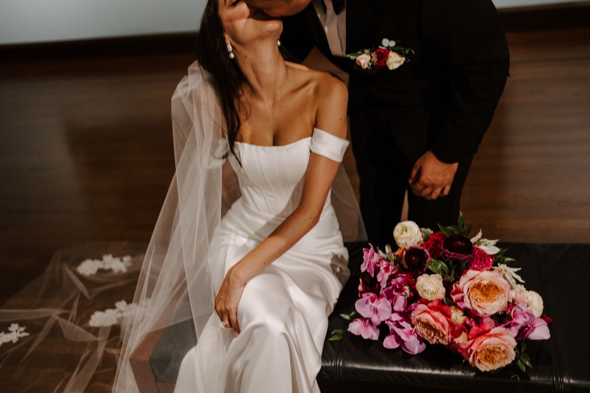 A bride in an elegant off-shoulder white wedding dress sits beside a colorful bouquet of pink and peach flowers.