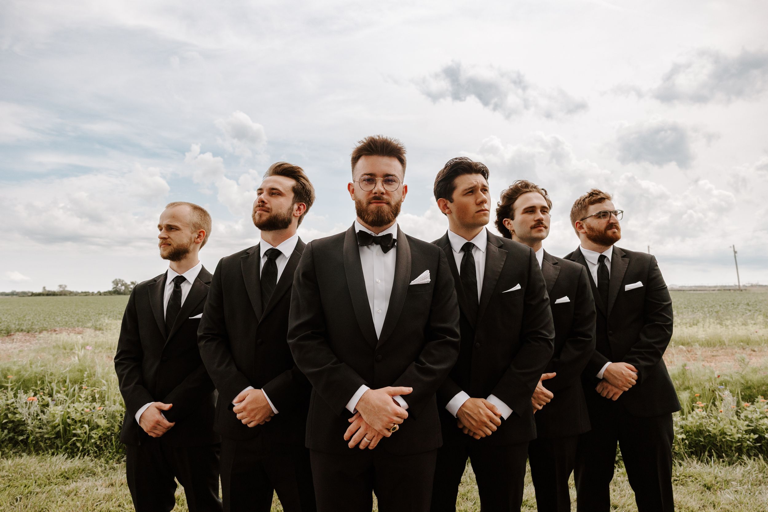 Groomsmen in black suits stand together in a line against a scenic outdoor landscape with cloudy skies.