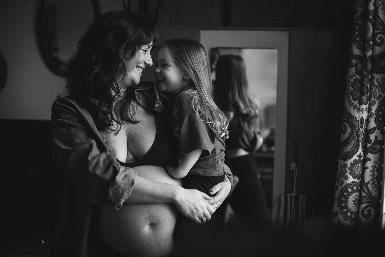 A tender black and white portrait of a parent and child embracing in a dimly lit room with a mirror in the background.