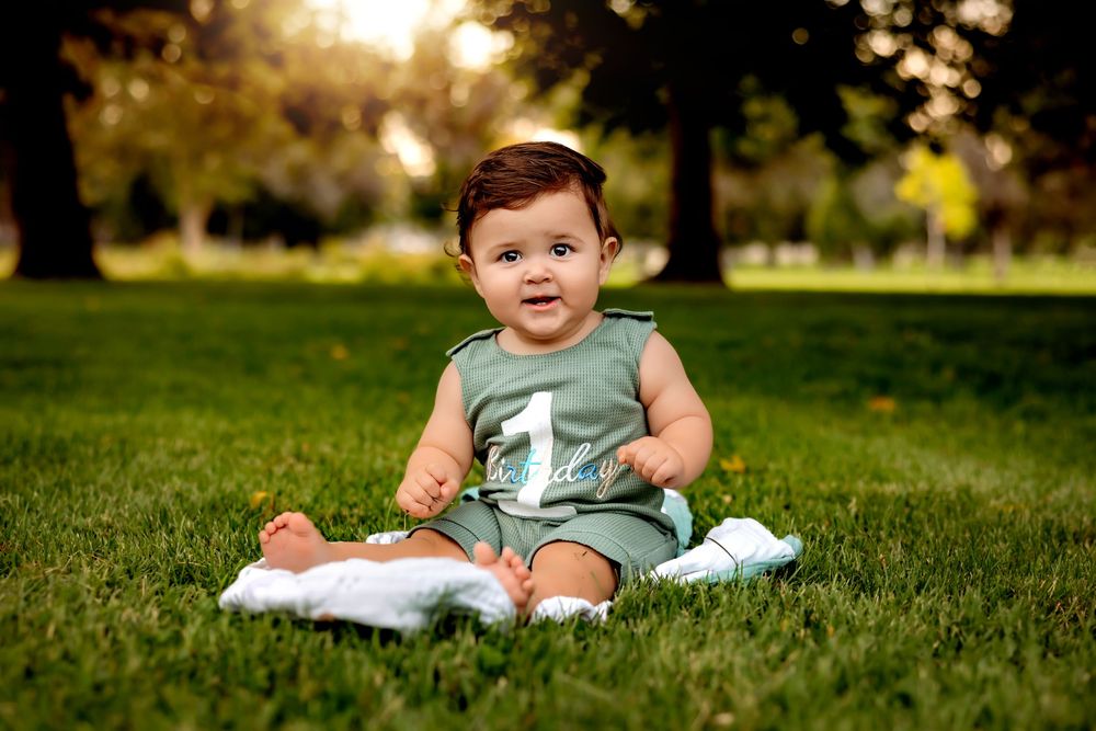 A baby in a sage green romper sits and plays on lush green grass in a sunny outdoor setting.