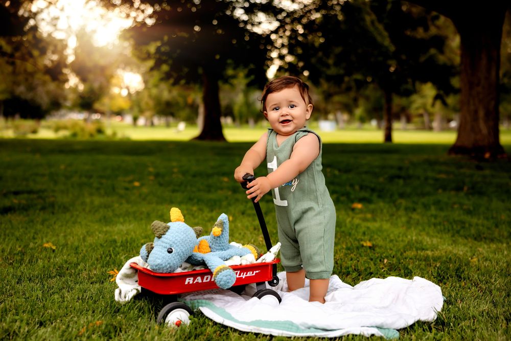 A baby sits on a white blanket in a sunny park with plush toys and stuffed animals.