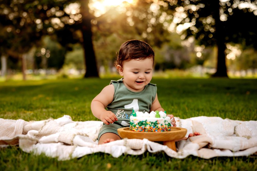 Baby in white outfit sits on blanket in grassy field during golden sunset hour.