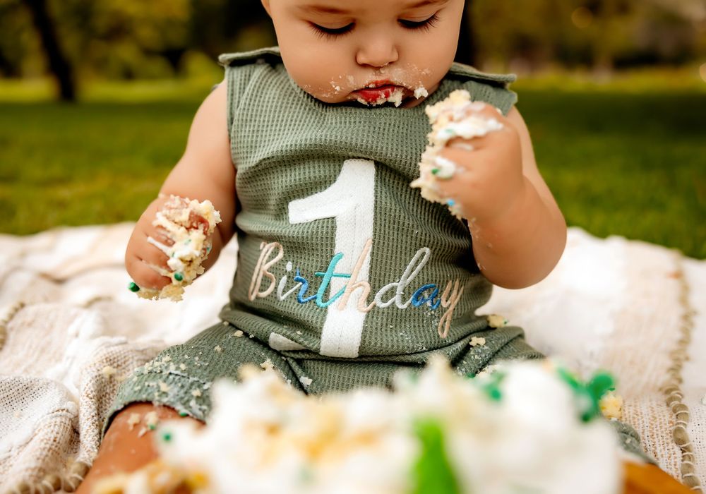 Baby in gray tank top enjoys birthday cake smash photo session outdoors on white blanket.