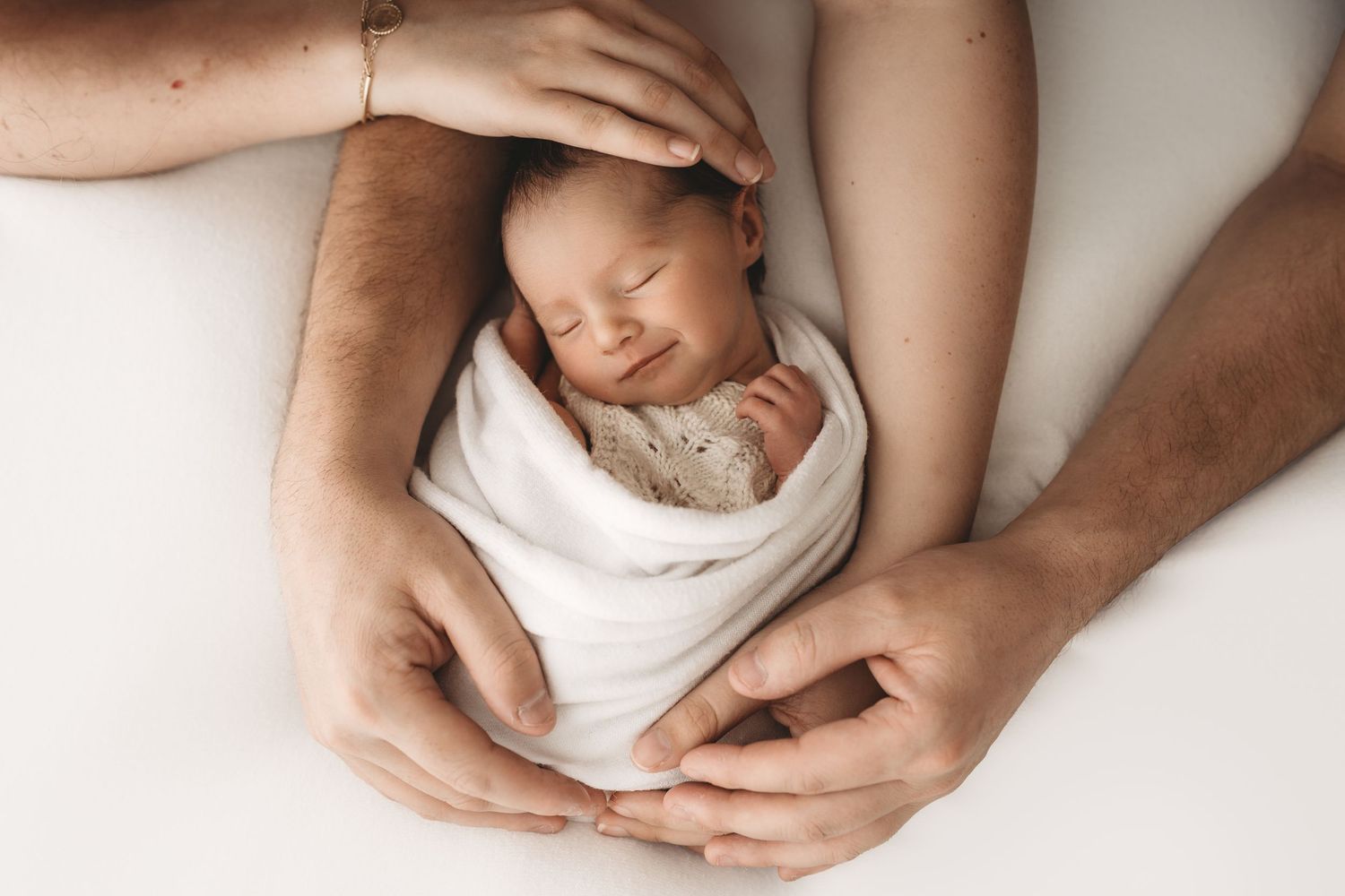 Parents cradling sleeping newborn baby wrapped in white swaddle, top view of family hands gently holding infant.
