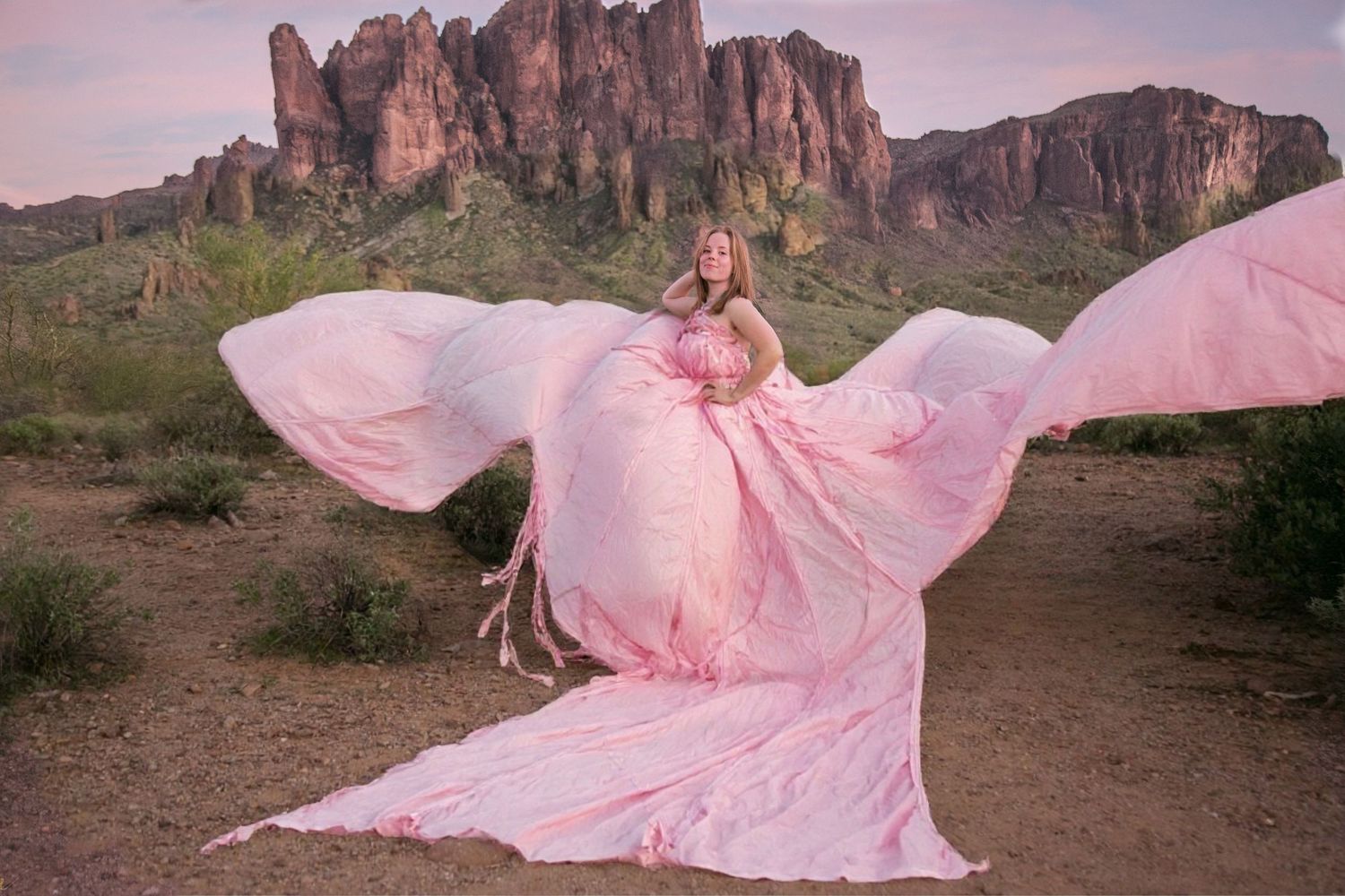 Woman in pink flowing gown poses in desert with dramatic rock formations at dusk.