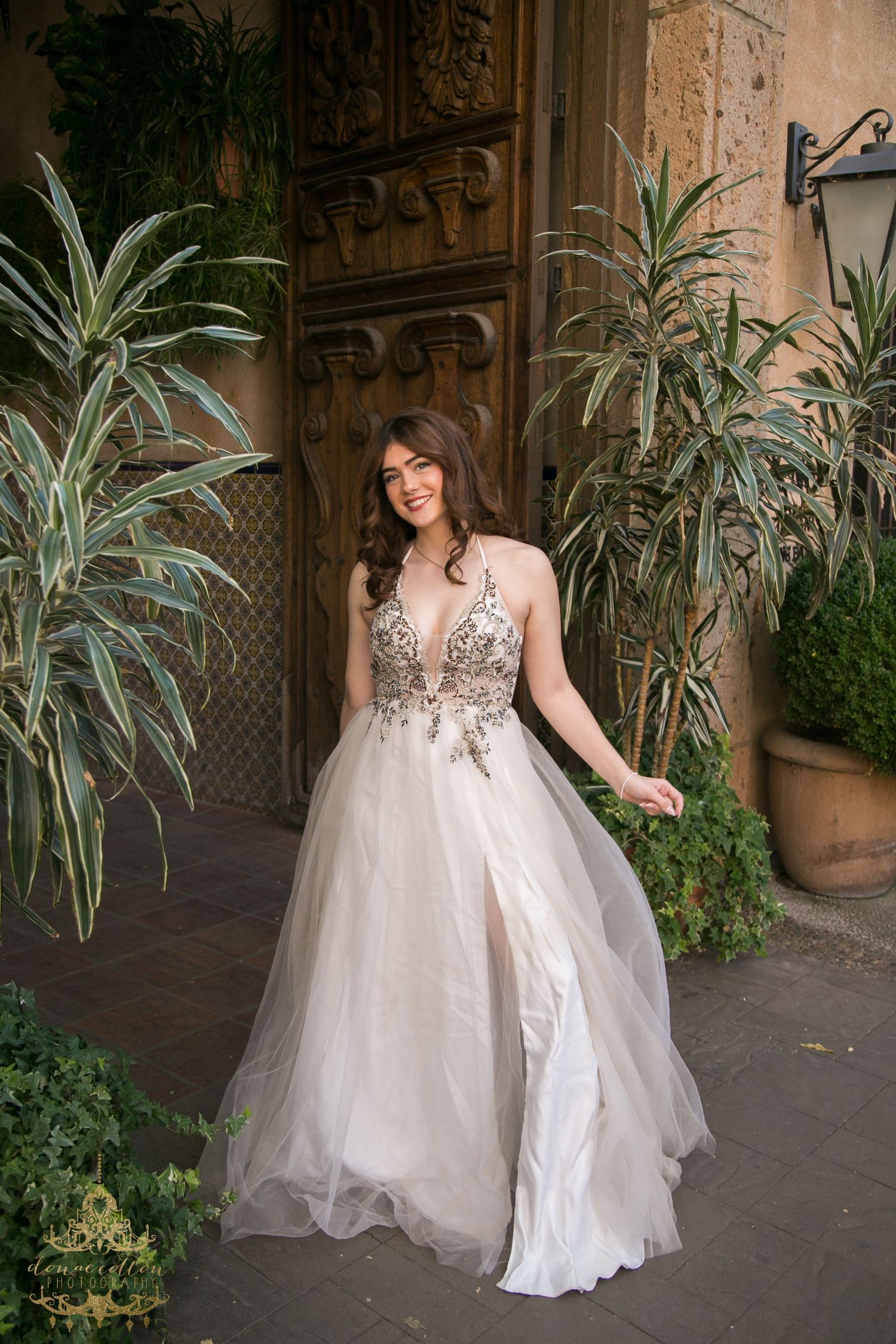 Bride in embroidered tulle wedding gown with slit poses by ornate wooden door and tropical plants.