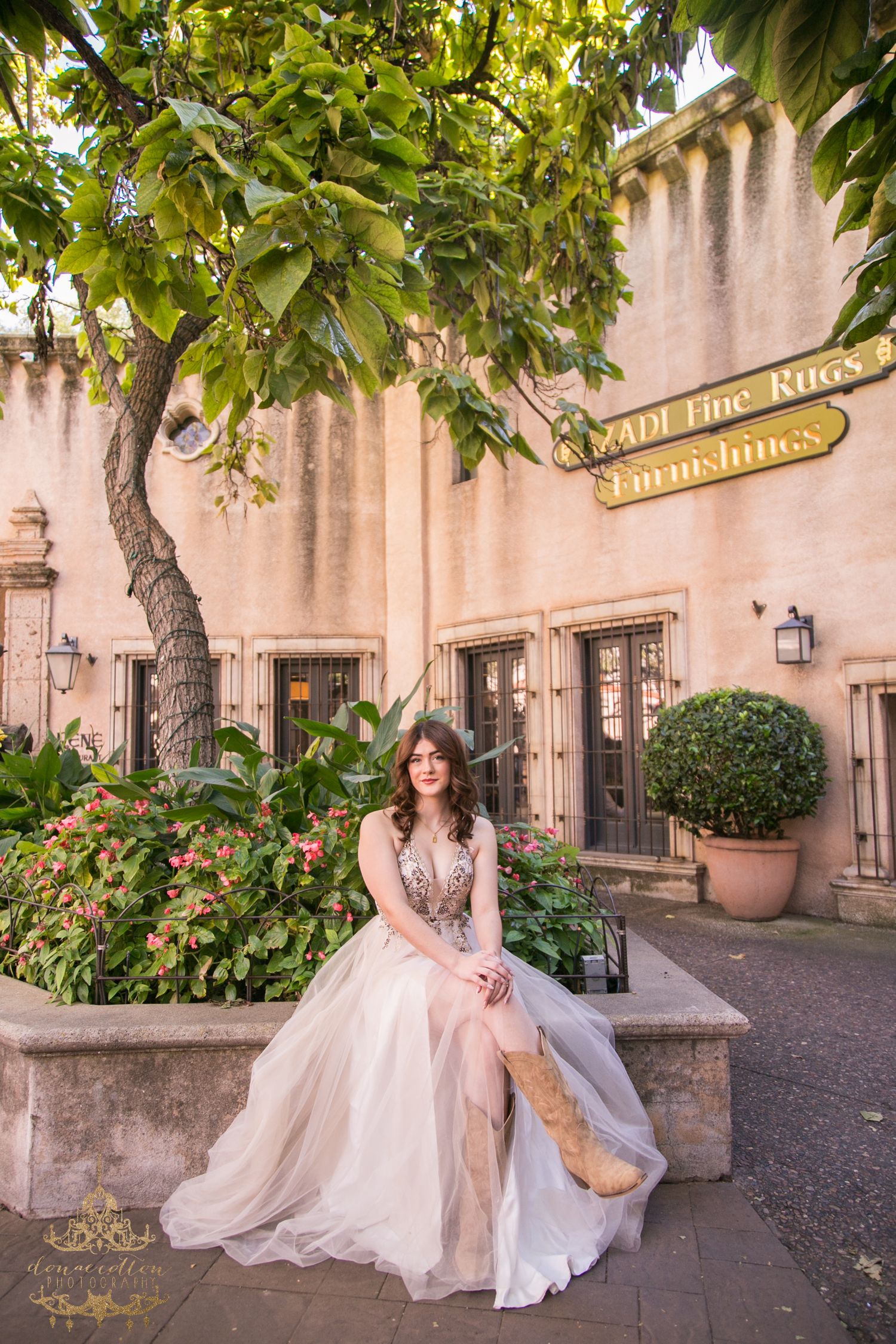 Bride in tulle gown with cowboy boots sits on garden bench outside rustic Scottsdale building.