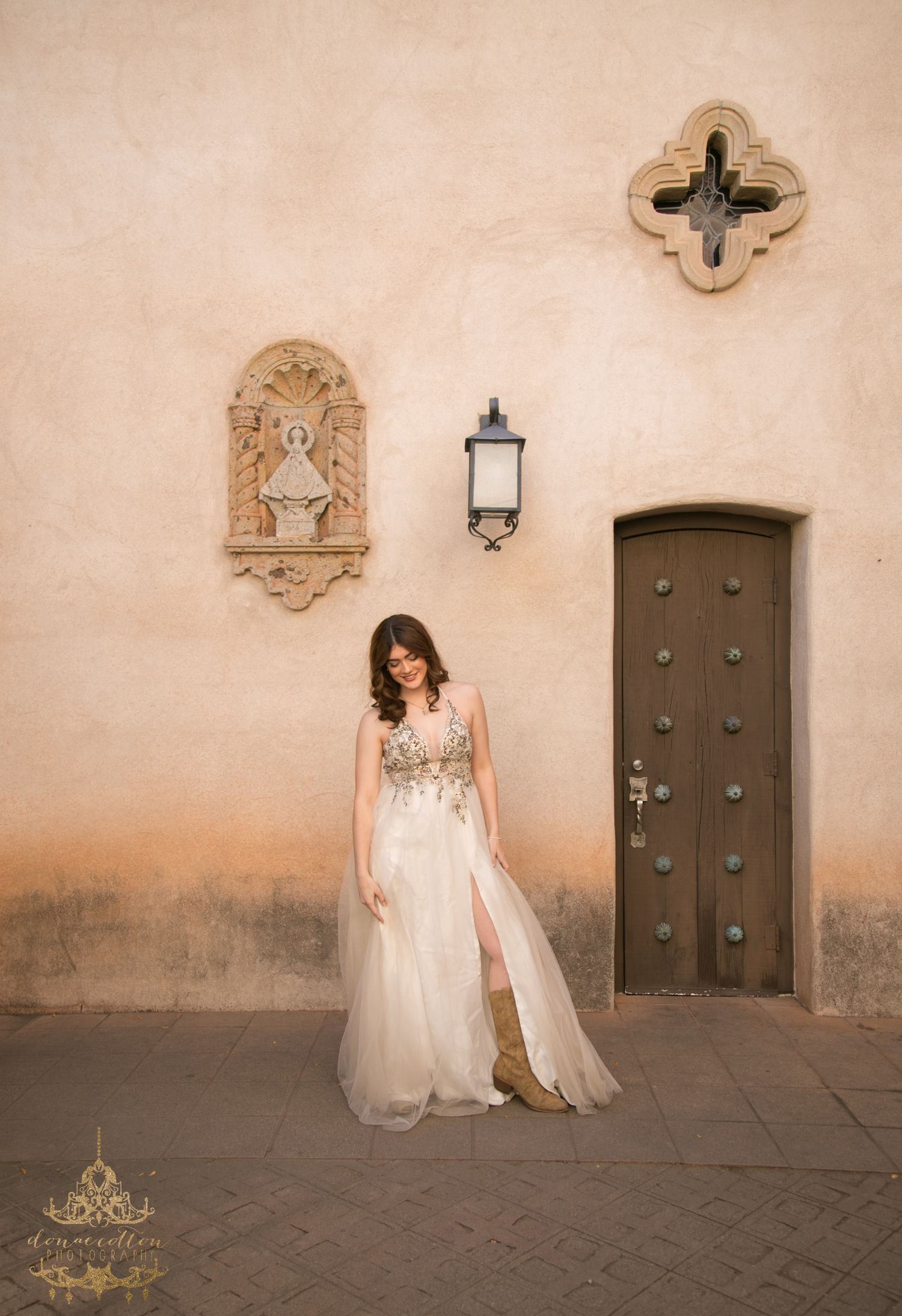 Bride in floral embroidered white gown poses before rustic adobe wall with wooden door.