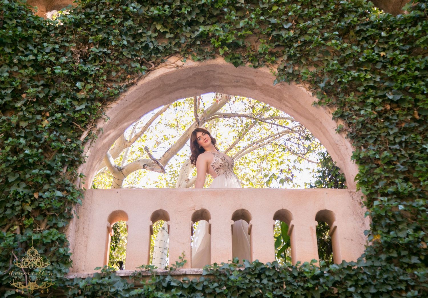 Bride in tulle gown poses gracefully inside ivy-covered arched window overlooking garden trees.