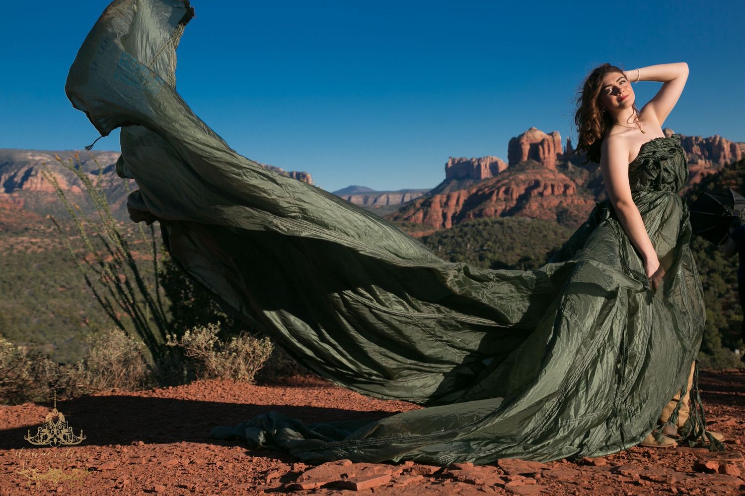 Woman in flowing olive green gown poses dramatically against Sedona red rock landscape with dress billowing in wind.
