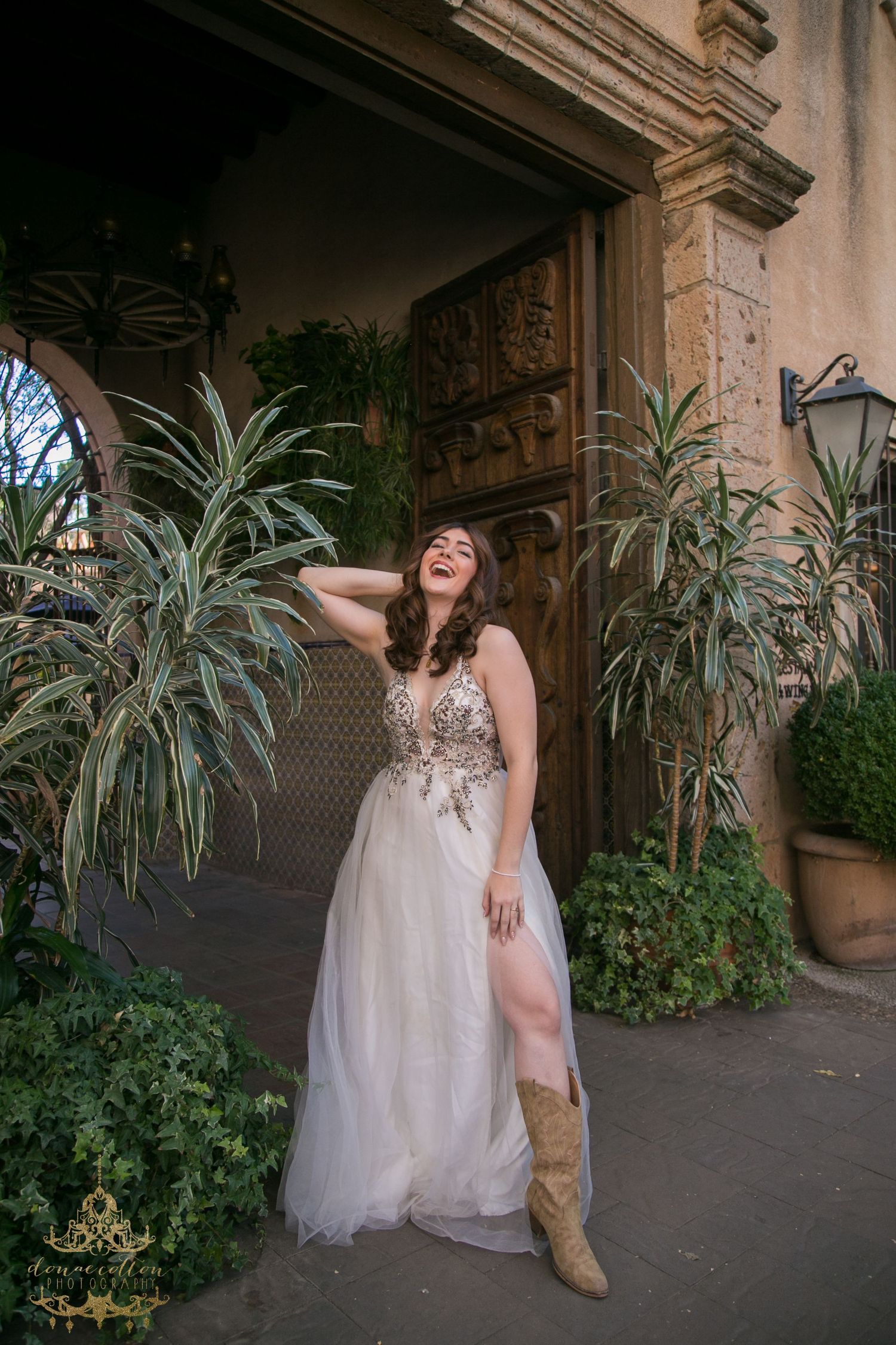 Laughing bride in embroidered tulle gown with cowboy boots poses by ornate door and plants.