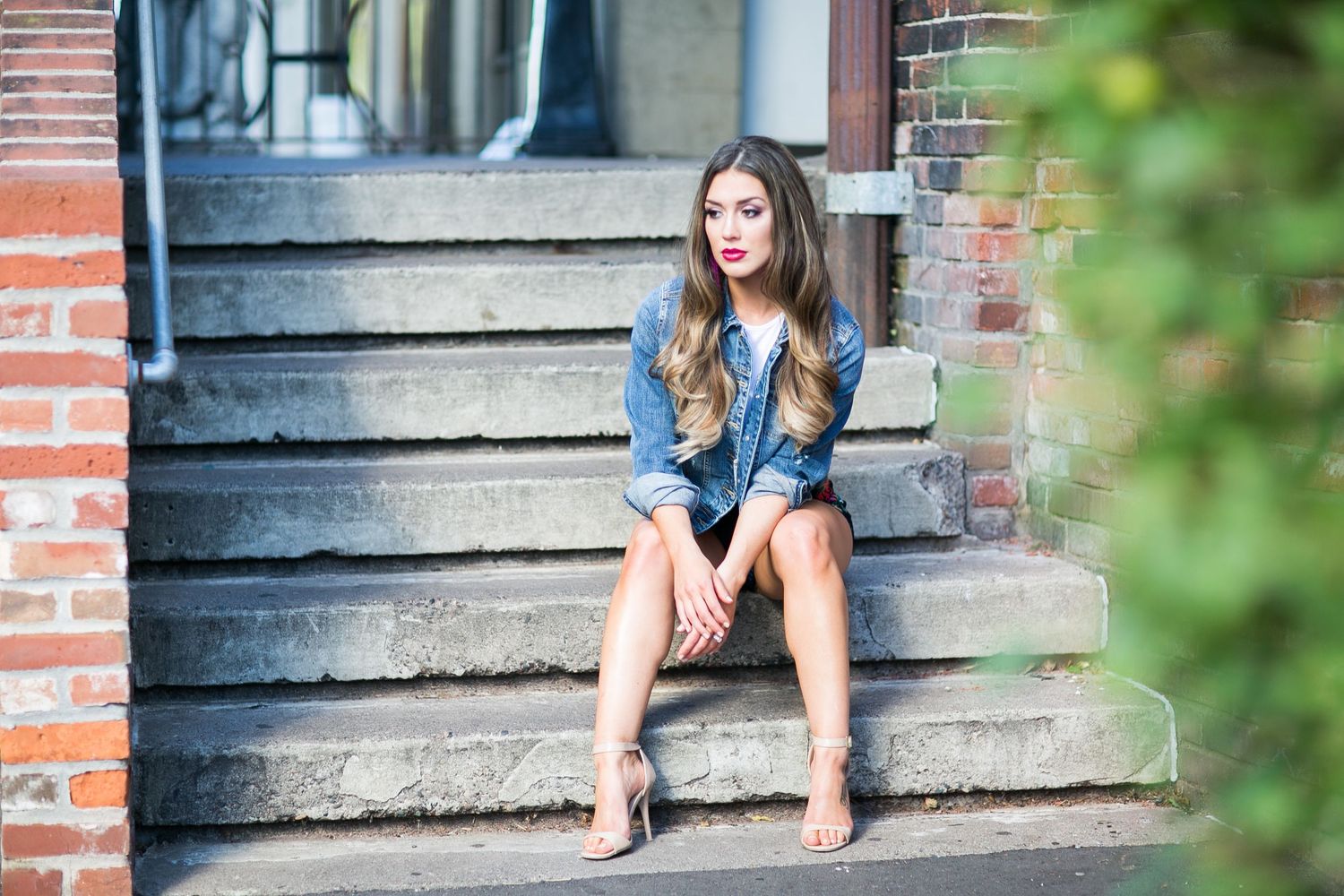 Young woman in denim jacket with red lips sits on stone steps in urban setting.