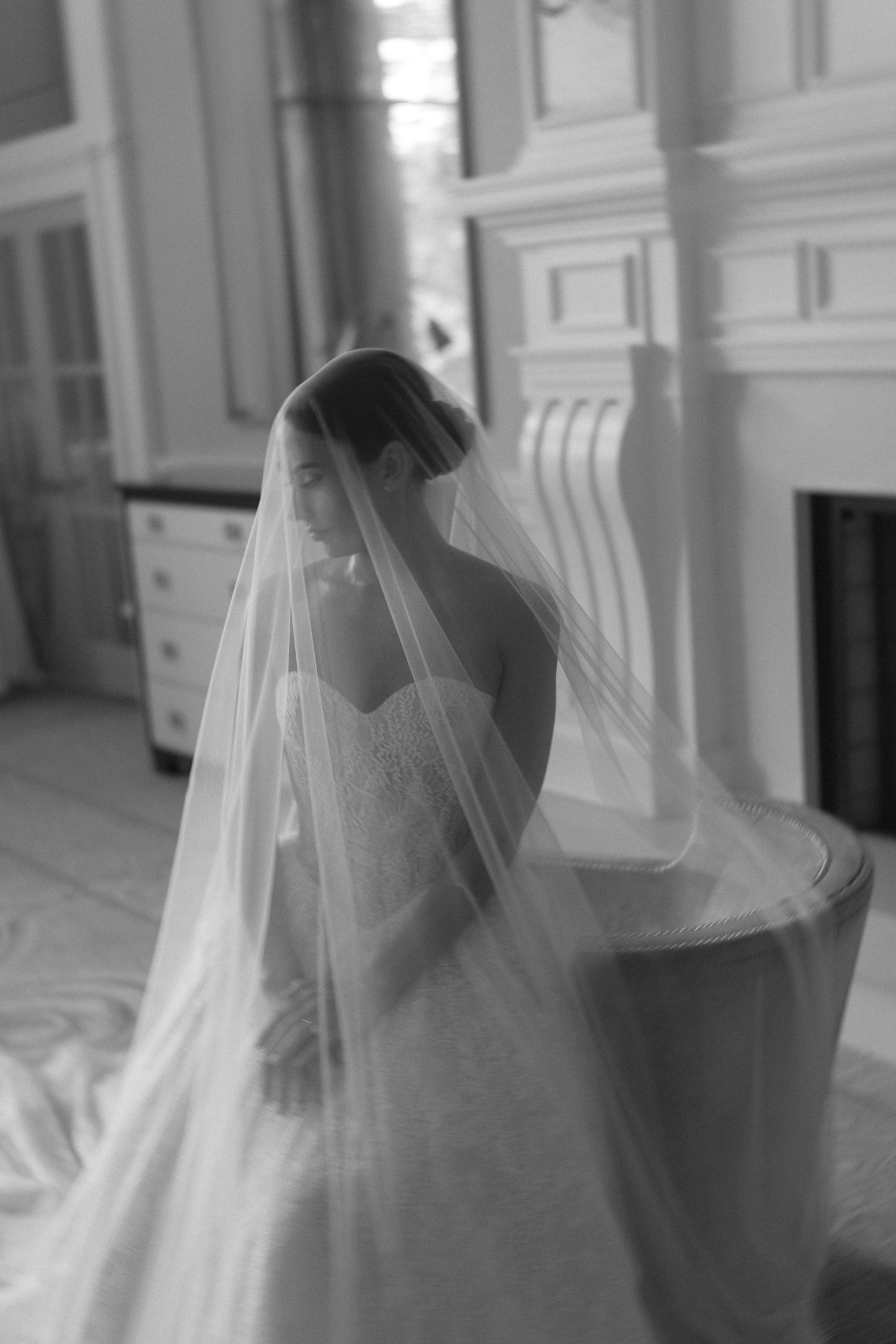 Elegant bride in a lace gown draped in a flowing veil in a black and white portrait.