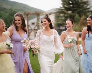 Wedding party in pastel bridesmaid dresses smiling and laughing together on a lawn with mountain backdrop.
