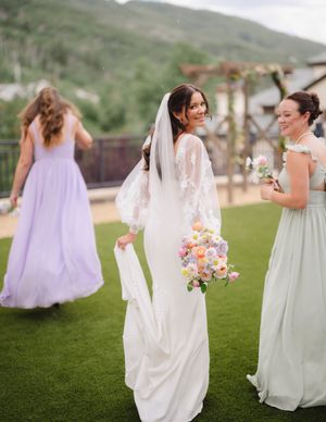Bride in long-sleeved lace gown with flowing veil poses with bridesmaids in pastel dresses on a lush outdoor lawn.