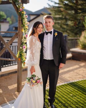 Newlyweds pose under a floral arch with mountain views and evergreen trees in the background.