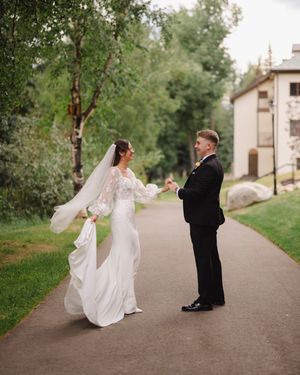A bride in a flowing white dress and groom in black suit share a playful moment on a garden path during their wedding day.