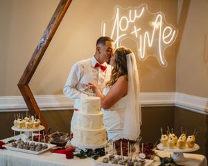 Dessert table with illuminated neon sign and elegant white cake display at wedding reception.