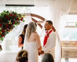 Wedding ceremony scene with red floral arrangements and white drapery.