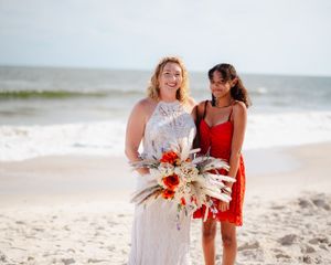 Two people in white and red dresses walk along a sunny beach holding floral bouquets by the ocean.