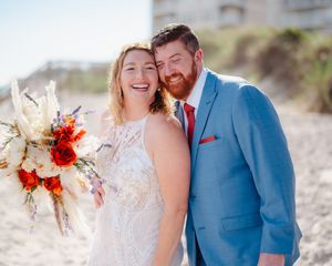 A romantic beach wedding couple stands together in blue and white attire with vibrant orange and white florals.