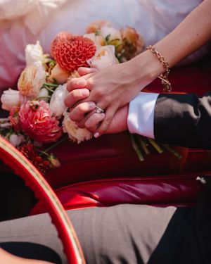 Close-up of joined hands with wedding rings resting on a bouquet of pink and coral flowers.