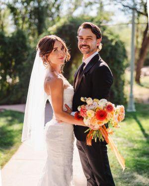 Wedding couple shares joyful moments with bright orange and white floral bouquets in a sunny garden.