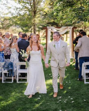 A wedding couple walks hand in hand across a grassy lawn surrounded by guests and white folding chairs on a sunny day.