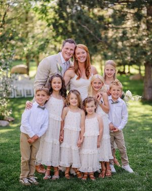 A group in white and beige outfits poses together outdoors on a sunny day in a grassy area with trees.