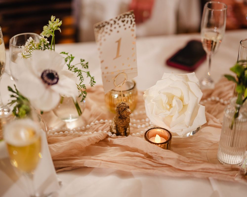 Elegant wedding reception table settings with white linens, glassware, and delicate floral arrangements in warm lighting.