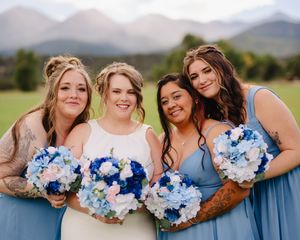 Wedding party holds blue and white floral bouquets in an outdoor setting with mountains in background.