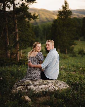 Couple sits together on a large rock surrounded by pine trees during sunset engagement photos.