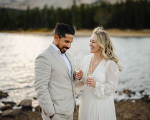 Couple in wedding attire sharing champagne by lakeside at sunset.