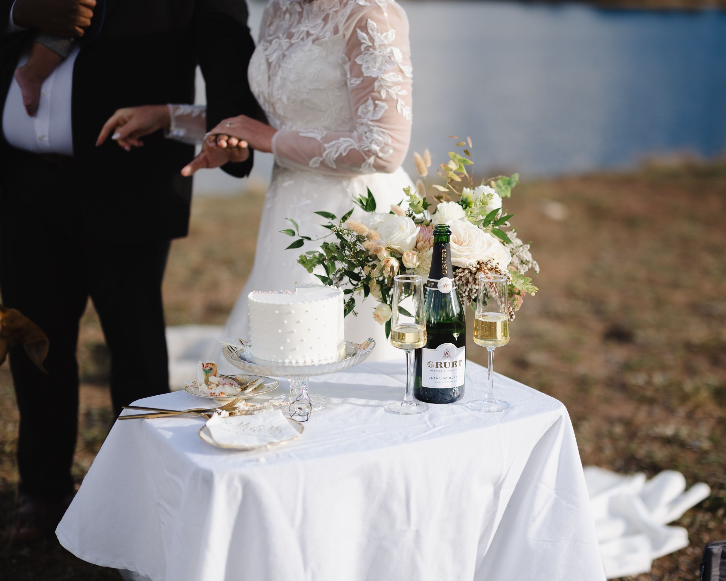 Elegant white wedding cake with floral decorations displayed on a round table at an outdoor reception.