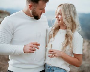 Couple in white outfits sharing drinks against mountain backdrop.