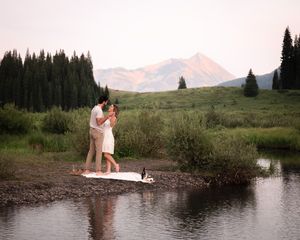 Couple stands on edge of mountain pond with evergreen forest and snow-capped peaks at sunset.
