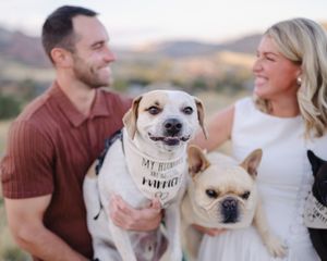 A couple in casual attire smiles while holding their small white dogs during an outdoor photo session.
