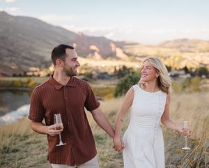 A couple holds wine glasses while enjoying a romantic sunset moment in a scenic mountain landscape.