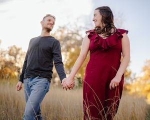 A couple holds hands while walking through tall autumn grass at sunset, with the person in a burgundy dress.