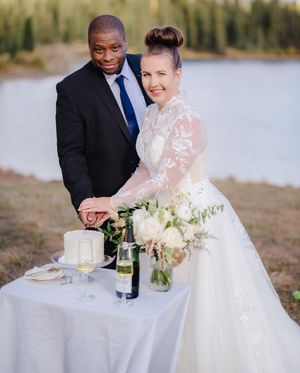 A couple in formal wedding attire shares a romantic moment at a lakeside table with wine and flowers.