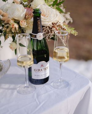 Elegant table setting with white roses in glass vases alongside champagne bottles and wine glasses on a white tablecloth.
