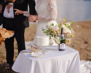 Elegant wedding cake display on white tablecloth with delicate floral decorations and wine bottles at an outdoor reception.