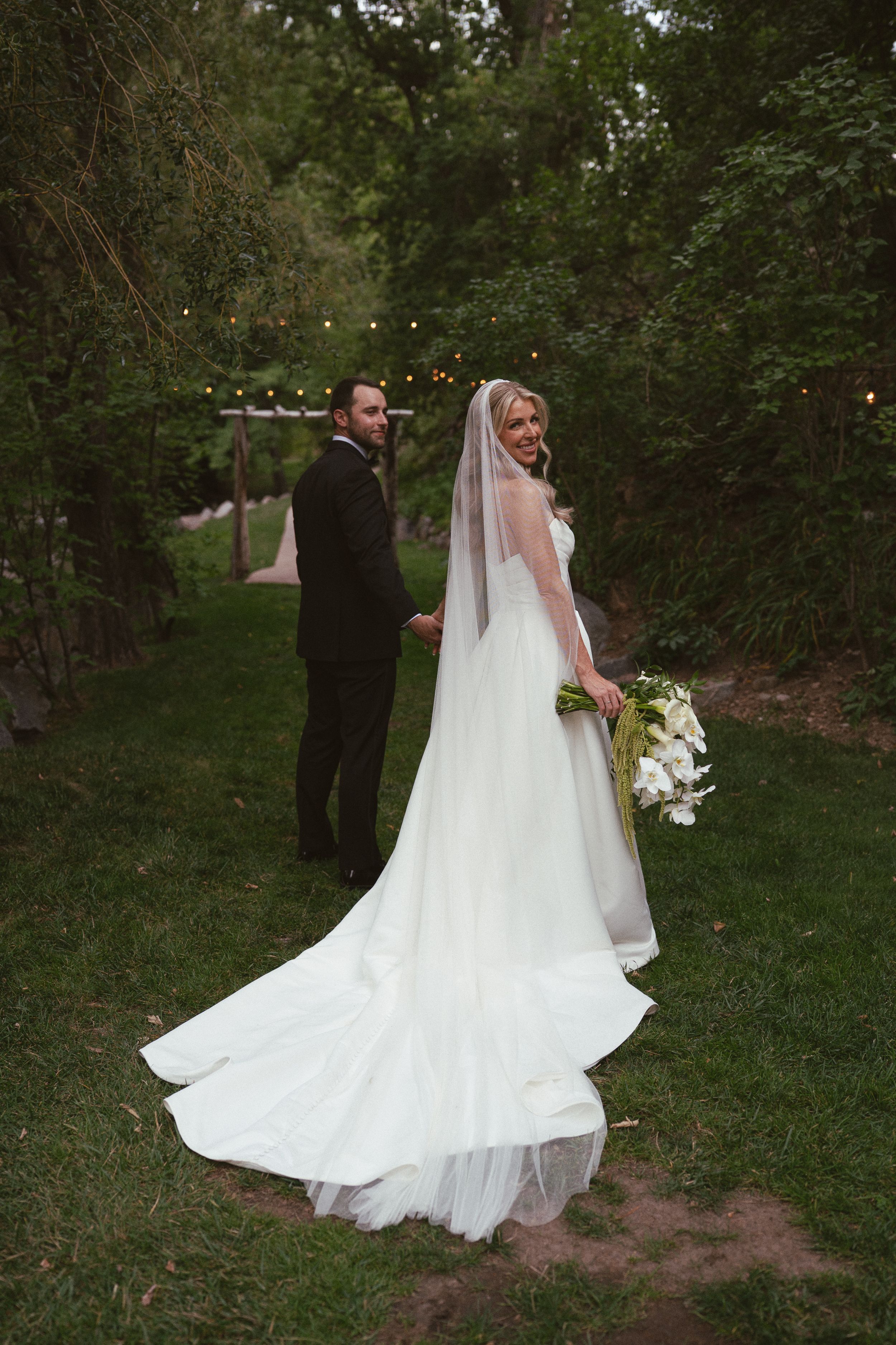 Bride in white gown with veil holds bouquet beside groom in dark suit, standing on green grass with string lights.