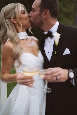 Bride and groom kissing while holding champagne glasses, wearing wedding attire with bow tie and white gown.