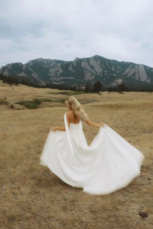 A blonde woman in a flowing white wedding dress twirls in a golden field with dramatic rocky mountains in the background.