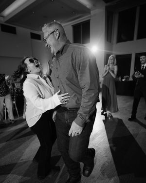 Black and white photo of a laughing couple dancing joyfully at a wedding reception with guests applauding in background.