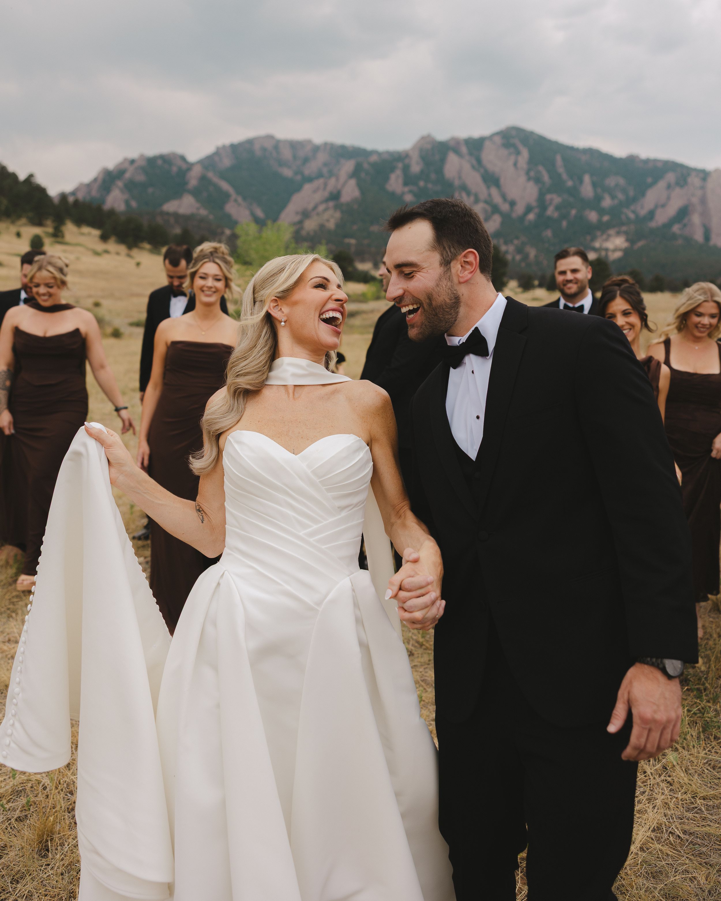 Bride and groom laugh while holding hands outdoors with Rocky Mountain Flatirons backdrop and wedding party in brown attire.